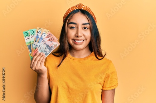 Young latin woman holding australian dollars banknotes looking positive and happy standing and smiling with a confident smile showing teeth