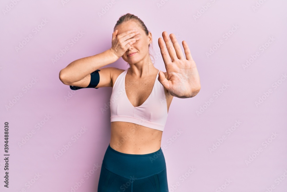 Beautiful blonde woman wearing sportswear and arm band covering eyes with hands and doing stop gesture with sad and fear expression. embarrassed and negative concept. Beautiful blonde woman wearing sportswear and arm band covering eyes with hands and doing stop gesture with sad and fear expression. embarrassed and negative concept.