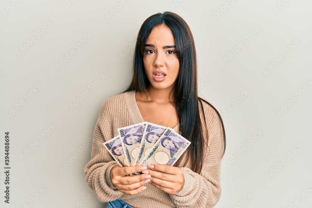 Young beautiful hispanic girl holding japanese yen banknotes clueless and confused expression. doubt concept. Young beautiful hispanic girl holding japanese yen banknotes clueless and confused expression. doubt concept.