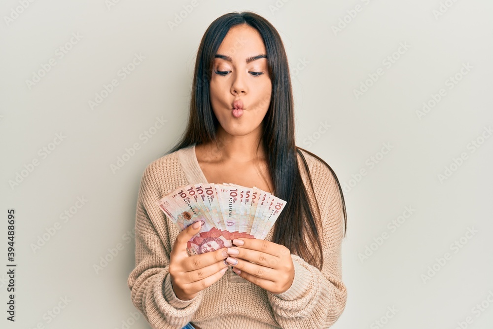 Young beautiful hispanic girl holding 10 colombian pesos banknotes making fish face with mouth and squinting eyes, crazy and comical. Young beautiful hispanic girl holding 10 colombian pesos banknotes making fish face with mouth and squinting eyes, crazy and comical.