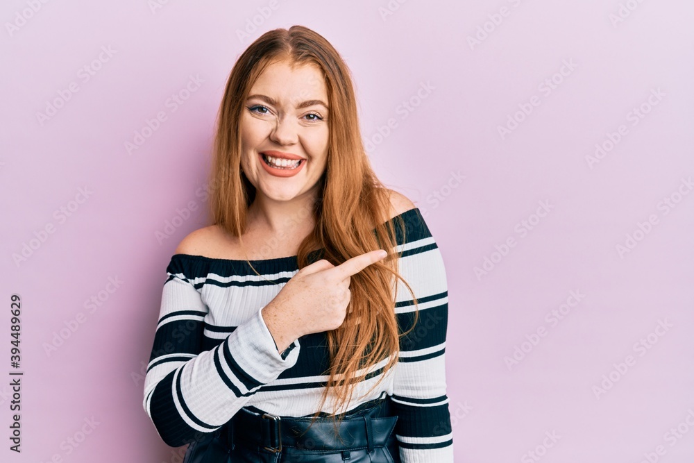 Young beautiful redhead woman wearing striped sweater over pink background smiling cheerful pointing with hand and finger up to the side Young beautiful redhead woman wearing striped sweater over pink background smiling cheerful pointing with hand and finger up to the side