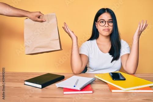 Tableau sur toile Young beautiful asian girl sitting on the table stuying getting take away food clueless and confused with open arms, no idea and doubtful face