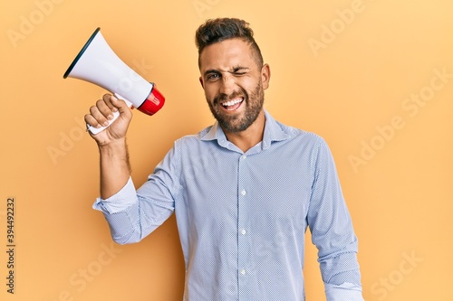 Handsome man with beard shouting through megaphone winking looking at the camera with sexy expression, cheerful and happy face.