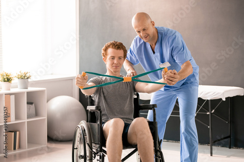Mature clinician in blue uniform supporting hand of young patient in wheelchair