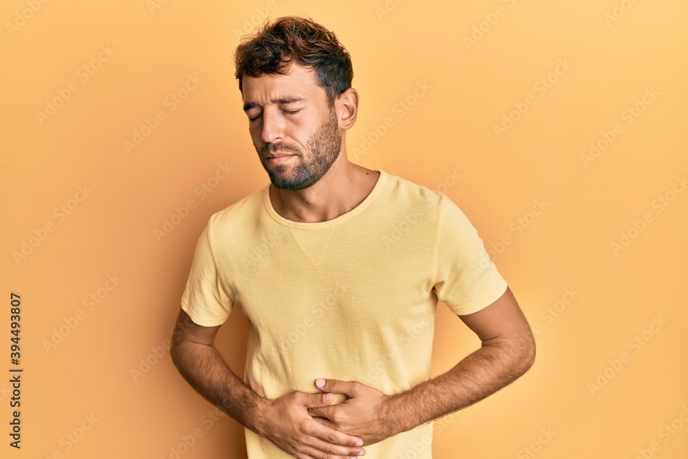 Handsome man with beard wearing casual yellow tshirt over yellow background with hand on stomach because indigestion, painful illness feeling unwell. ache concept. Handsome man with beard wearing casual yellow tshirt over yellow background with hand on stomach because indigestion, painful illness feeling unwell. ache concept.