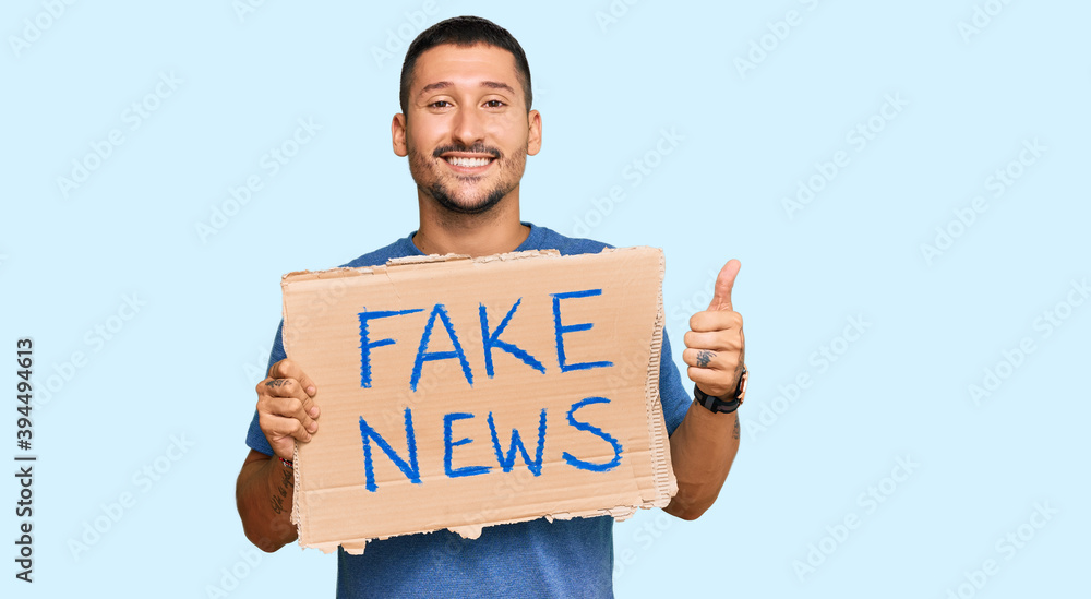 Handsome man with tattoos holding fake news banner smiling happy and positive, thumb up doing excellent and approval sign Handsome man with tattoos holding fake news banner smiling happy and positive, thumb up doing excellent and approval sign