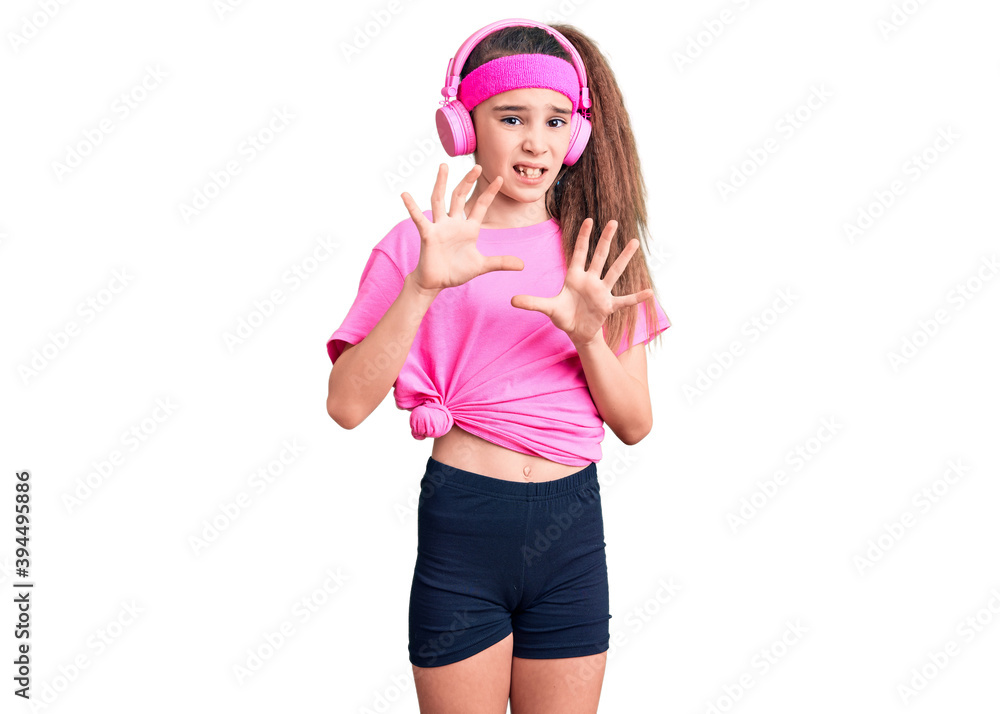 Cute hispanic child girl wearing gym clothes and using headphones disgusted expression, displeased and fearful doing disgust face because aversion reaction. with hands raised Cute hispanic child girl wearing gym clothes and using headphones disgusted expression, displeased and fearful doing disgust face because aversion reaction. with hands raised