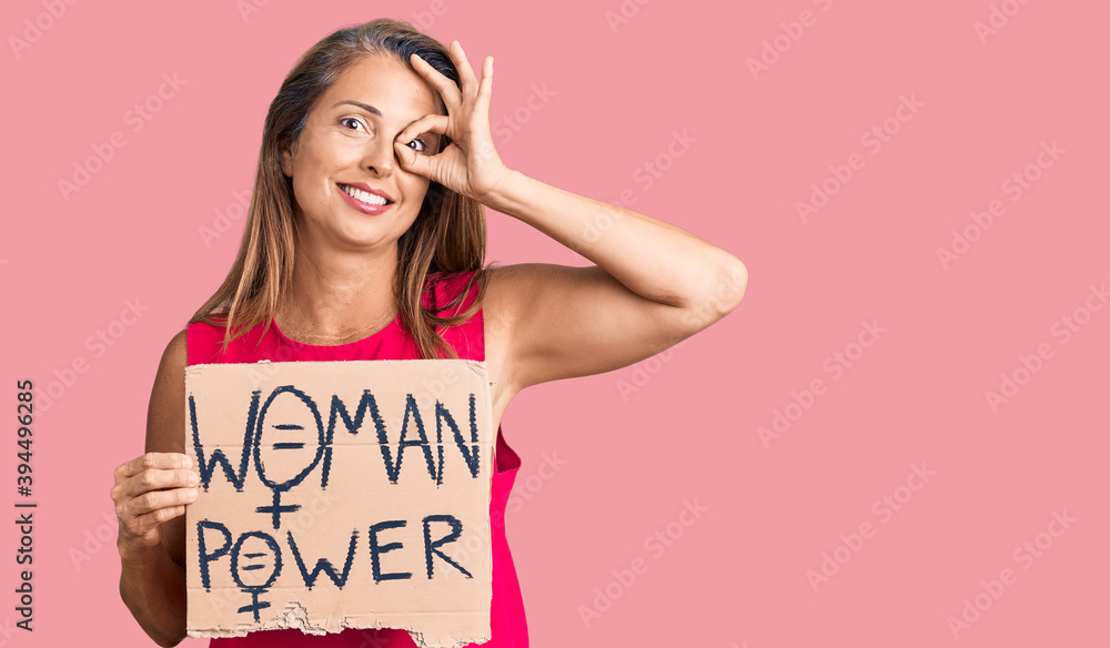 Middle age hispanic woman holding woman power banner smiling happy doing ok sign with hand on eye looking through fingers Middle age hispanic woman holding woman power banner smiling happy doing ok sign with hand on eye looking through fingers
