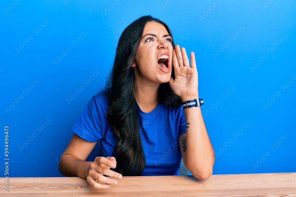 Beautiful brunette young woman wearing casual clothes sitting on the table shouting and screaming loud to side with hand on mouth. communication concept. Beautiful brunette young woman wearing casual clothes sitting on the table shouting and screaming loud to side with hand on mouth. communication concept.