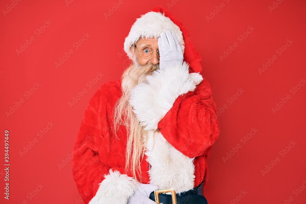 Old senior man with grey hair and long beard wearing traditional santa claus costume covering one eye with hand, confident smile on face and surprise emotion. Old senior man with grey hair and long beard wearing traditional santa claus costume covering one eye with hand, confident smile on face and surprise emotion.