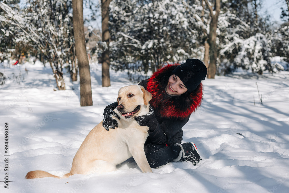Young woman sits on a snow in the park with her dog labrador retriever hugging. Female and her pet having fun outdoors on the winter bright sunny day. Friendship and togetherness concept. Young woman sits on a snow in the park with her dog labrador retriever hugging. Female and her pet having fun outdoors on the winter bright sunny day. Friendship and togetherness concept.