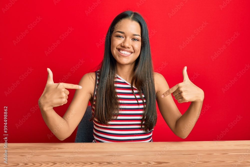 Beautiful hispanic woman wearing casual clothes sitting on the table looking confident with smile on face, pointing oneself with fingers proud and happy. Beautiful hispanic woman wearing casual clothes sitting on the table looking confident with smile on face, pointing oneself with fingers proud and happy.