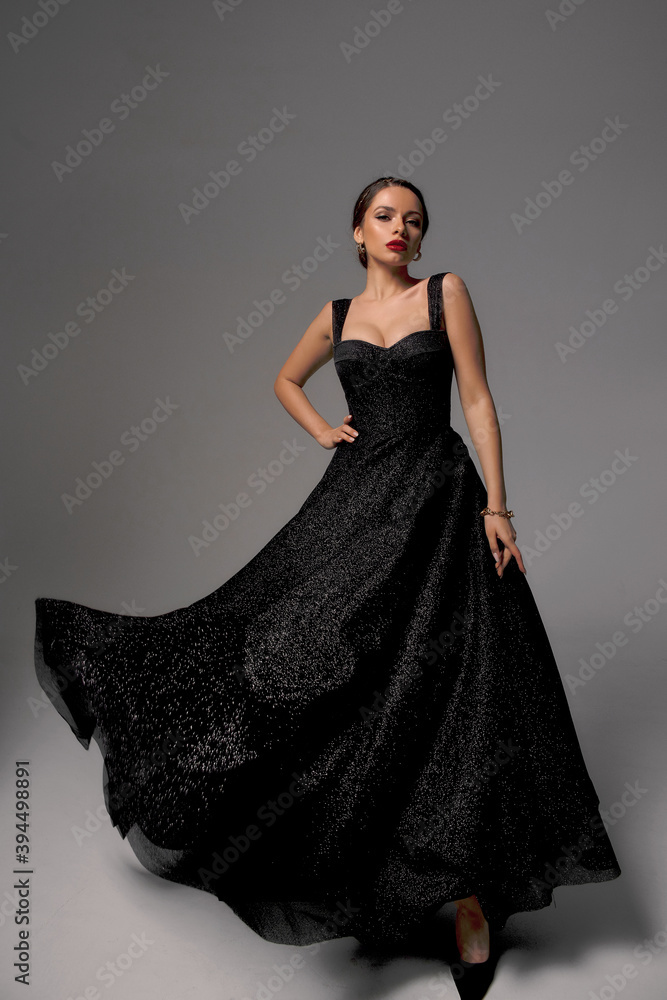 Full length studio fashion portrait. Elegant woman in black shiny evening dress. Dynamic shot. Fashion model with makeup and hairstyle. Caucasian brunette tall lady