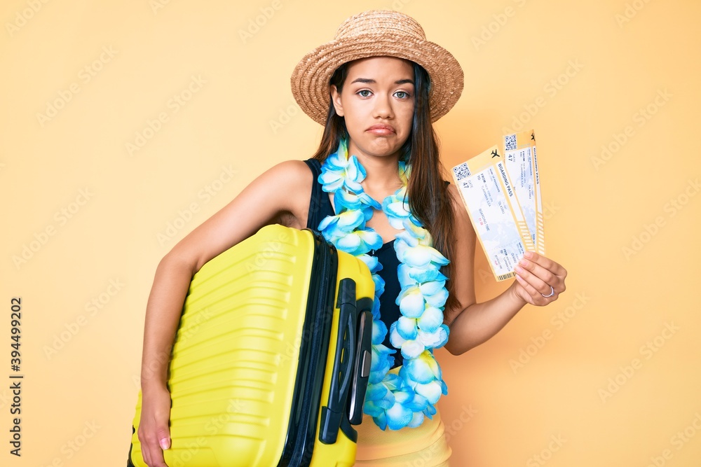 Young beautiful latin girl wearing summer hat and hawaiian lei holding cabin bag and boarding pass depressed and worry for distress, crying angry and afraid. sad expression.