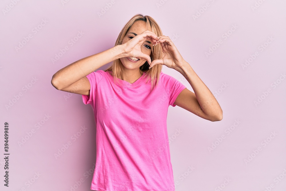Beautiful blonde woman wearing casual pink tshirt doing heart shape with hand and fingers smiling looking through sign Beautiful blonde woman wearing casual pink tshirt doing heart shape with hand and fingers smiling looking through sign