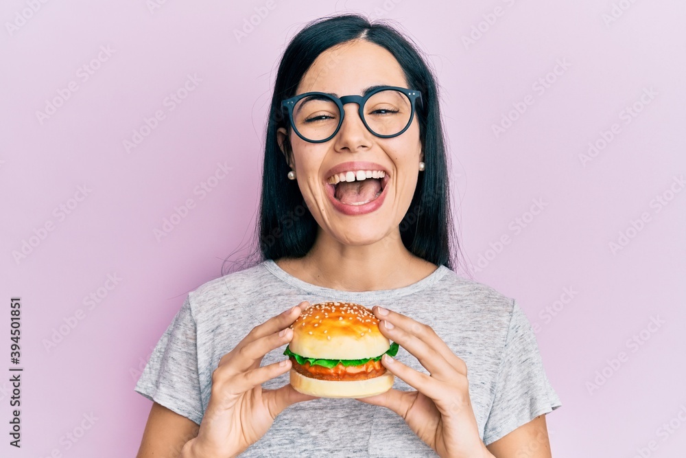 Beautiful young woman eating tasty hamburger smiling and laughing hard out loud because funny crazy joke.