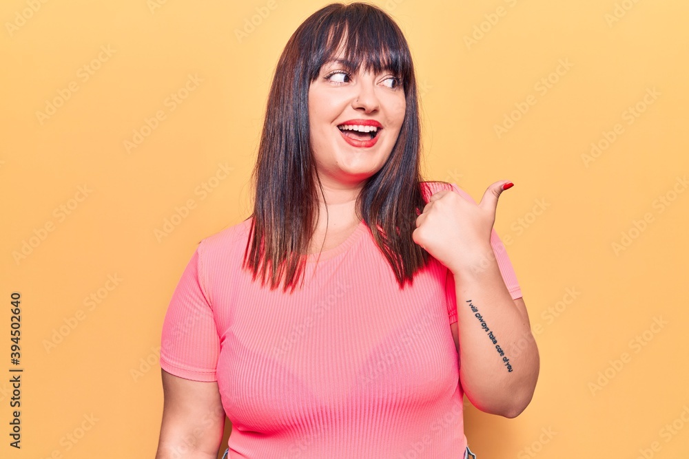 Young plus size woman wearing casual clothes smiling with happy face looking and pointing to the side with thumb up. Young plus size woman wearing casual clothes smiling with happy face looking and pointing to the side with thumb up.