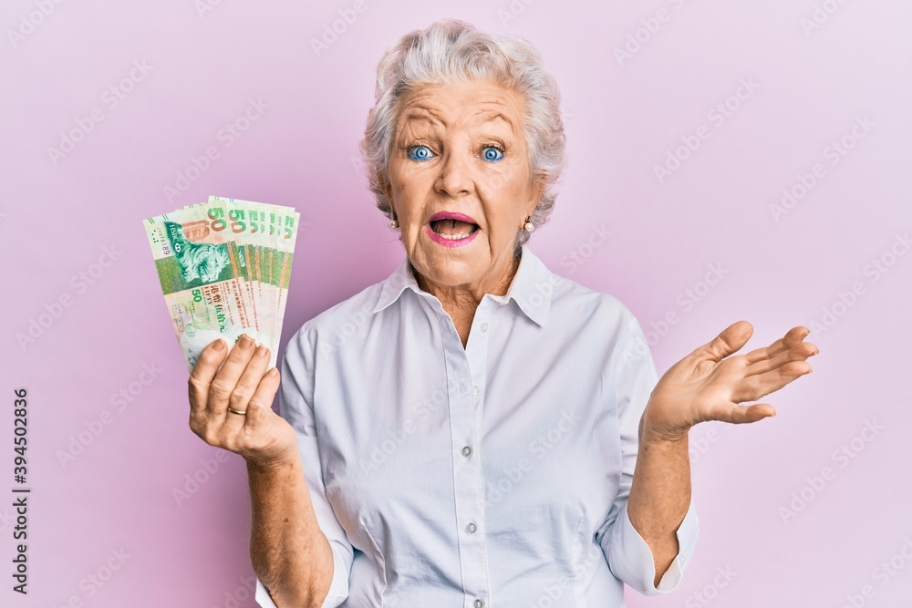 Senior grey-haired woman holding hong kong 50 dollars banknotes celebrating achievement with happy smile and winner expression with raised hand Senior grey-haired woman holding hong kong 50 dollars banknotes celebrating achievement with happy smile and winner expression with raised hand
