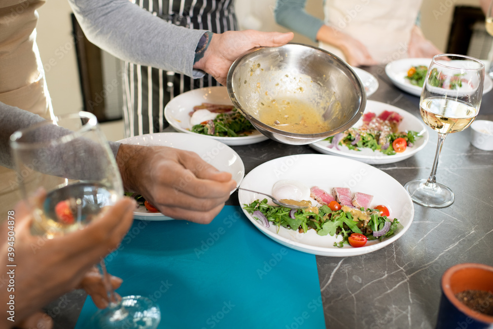 Hands of mature man putting sauce from metallic bowl on plate with salad Hands of mature man putting sauce from metallic bowl on plate with salad
