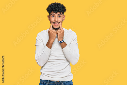 Young arab man wearing casual winter sweater smiling with open mouth, fingers pointing and forcing cheerful smile © Krakenimages.com