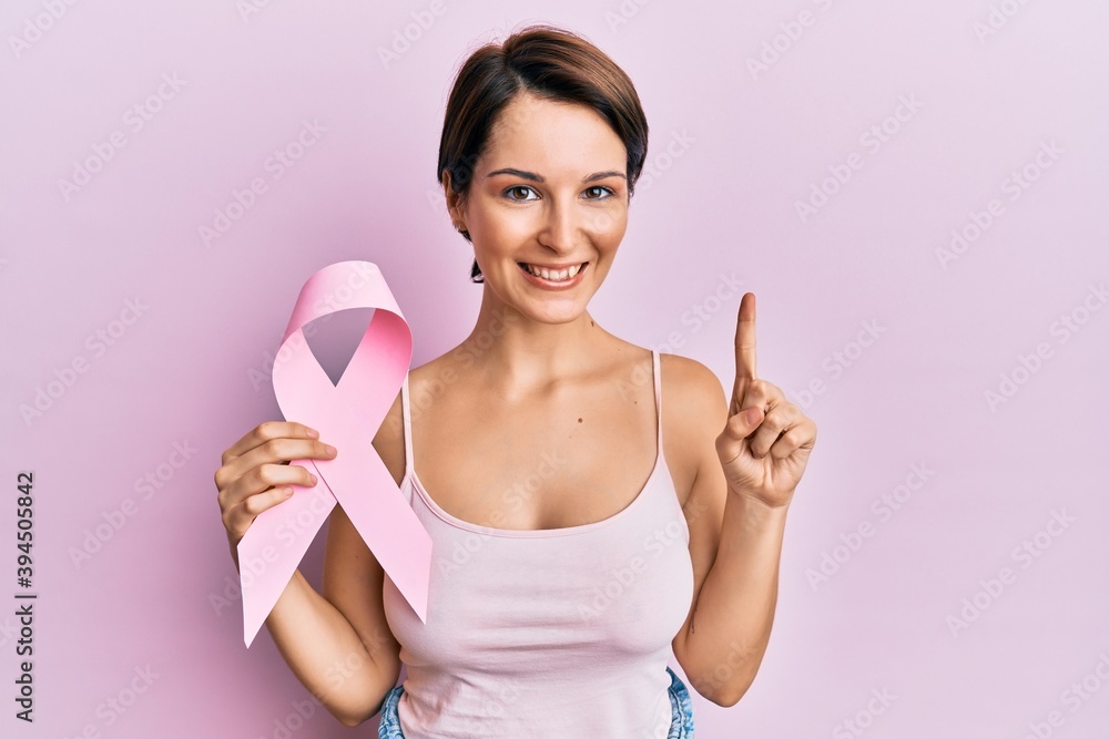 Young brunette woman with short hair holding pink cancer ribbon smiling with an idea or question pointing finger with happy face, number one Young brunette woman with short hair holding pink cancer ribbon smiling with an idea or question pointing finger with happy face, number one