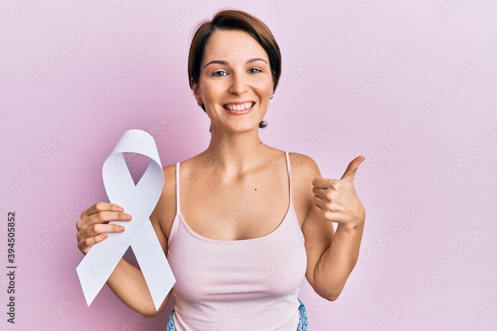 Young brunette woman with short hair holding cancer awareness white ribbon smiling happy and positive, thumb up doing excellent and approval sign Young brunette woman with short hair holding cancer awareness white ribbon smiling happy and positive, thumb up doing excellent and approval sign