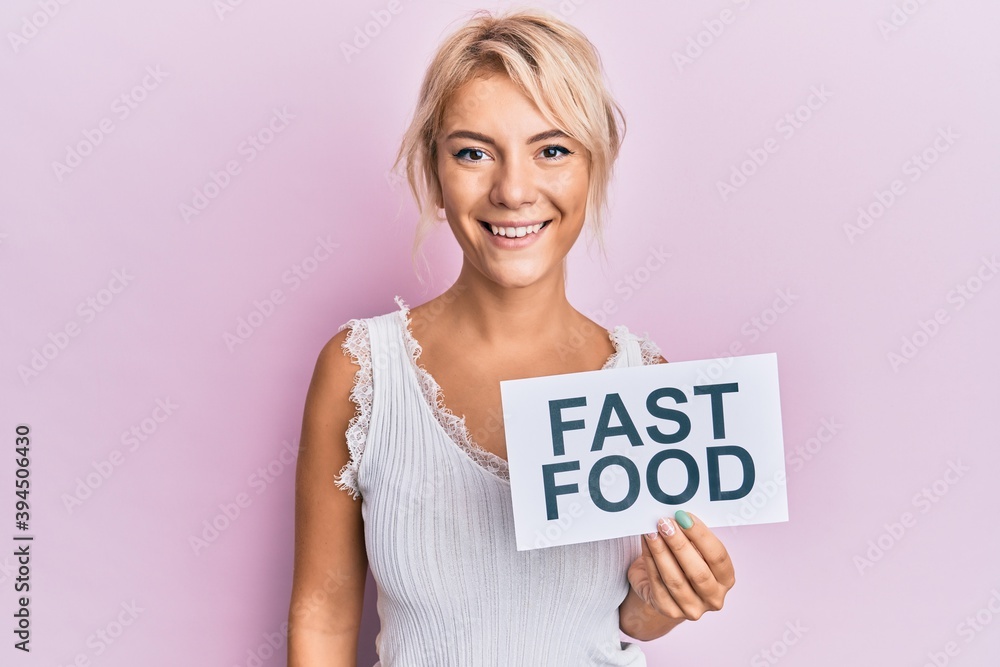 Young blonde girl eating fast food pizza and burger looking positive and happy standing and smiling with a confident smile showing teeth Young blonde girl eating fast food pizza and burger looking positive and happy standing and smiling with a confident smile showing teeth