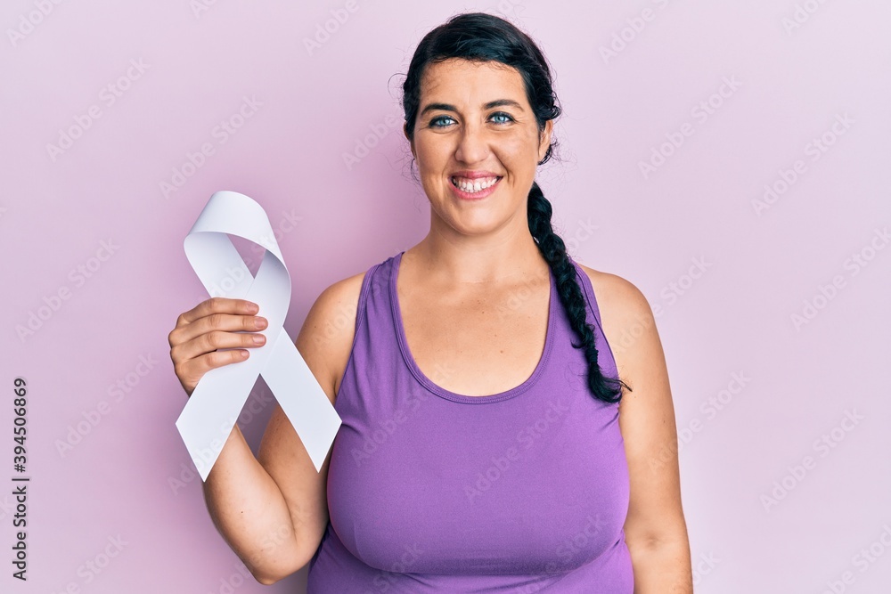 Plus size brunette woman holding white ribbon looking positive and happy standing and smiling with a confident smile showing teeth Plus size brunette woman holding white ribbon looking positive and happy standing and smiling with a confident smile showing teeth