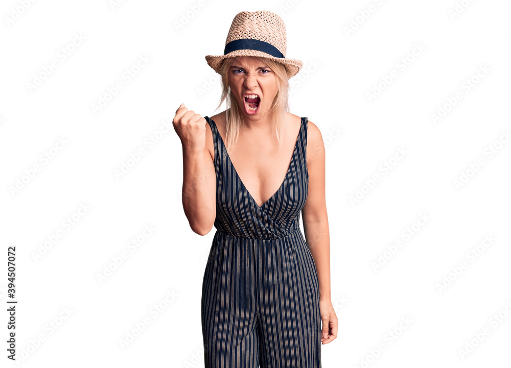 Young beautiful blonde woman wearing summer hat and dress angry and mad raising fist frustrated and furious while shouting with anger. rage and aggressive concept. Young beautiful blonde woman wearing summer hat and dress angry and mad raising fist frustrated and furious while shouting with anger. rage and aggressive concept.