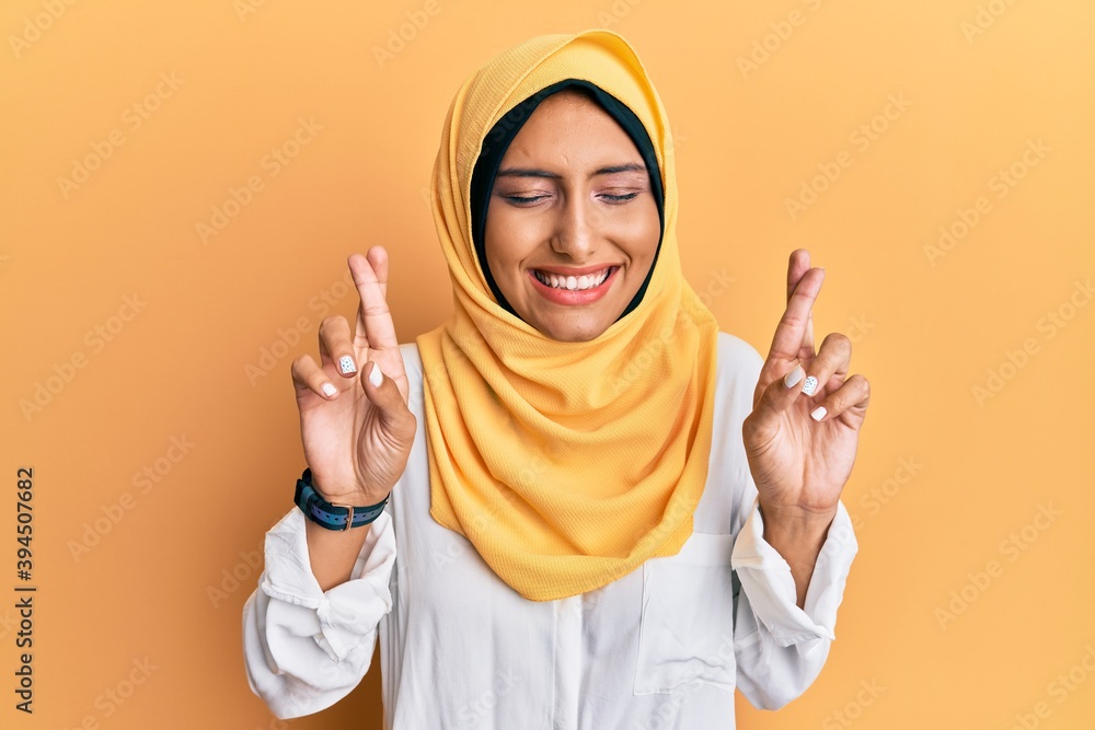 Young brunette arab woman wearing traditional islamic hijab scarf gesturing finger crossed smiling with hope and eyes closed. luck and superstitious concept. Young brunette arab woman wearing traditional islamic hijab scarf gesturing finger crossed smiling with hope and eyes closed. luck and superstitious concept.