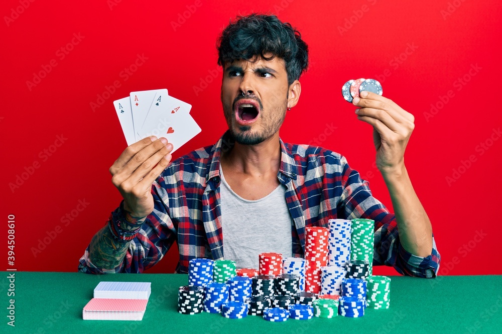 Young hispanic man playing poker holding cards and casino chips angry and mad screaming frustrated and furious, shouting with anger. rage and aggressive concept. Young hispanic man playing poker holding cards and casino chips angry and mad screaming frustrated and furious, shouting with anger. rage and aggressive concept.