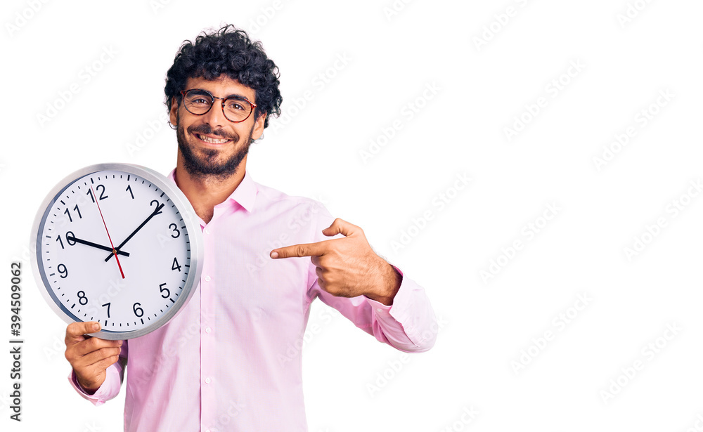 Handsome young man with curly hair and bear holding big clock smiling happy pointing with hand and finger Handsome young man with curly hair and bear holding big clock smiling happy pointing with hand and finger