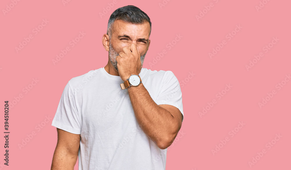 Middle age handsome man wearing casual white tshirt smelling something stinky and disgusting, intolerable smell, holding breath with fingers on nose. bad smell Middle age handsome man wearing casual white tshirt smelling something stinky and disgusting, intolerable smell, holding breath with fingers on nose. bad smell
