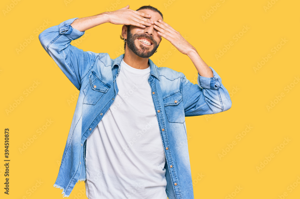 Attractive man with long hair and beard wearing casual denim jacket covering eyes with hands smiling cheerful and funny. blind concept. Attractive man with long hair and beard wearing casual denim jacket covering eyes with hands smiling cheerful and funny. blind concept.
