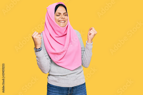 Young caucasian woman wearing traditional islamic hijab scarf very happy and excited doing winner gesture with arms raised, smiling and screaming for success. celebration concept. © Krakenimages.com