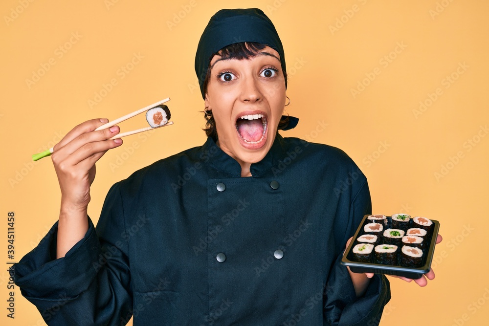 Beautiful brunettte woman chef holding sushi using chopsticks celebrating crazy and amazed for success with open eyes screaming excited. Beautiful brunettte woman chef holding sushi using chopsticks celebrating crazy and amazed for success with open eyes screaming excited.