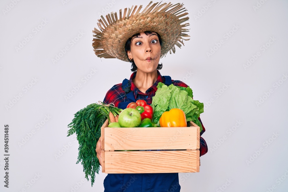 Beautiful brunettte woman wearing farmer clothes holding vegetables making fish face with mouth and squinting eyes, crazy and comical.