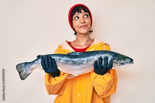 Foto Beautiful brunettte fisher woman wearing raincoat holding fresh salmon smiling looking to the side and staring away thinking
