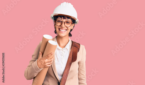 Young brunette woman with short hair wearing safety helmet holding blueprints looking positive and happy standing and smiling with a confident smile showing teeth