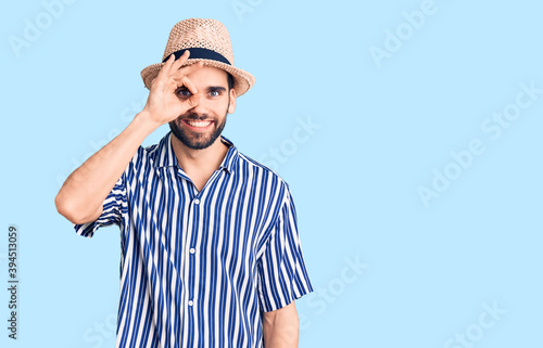 Young handsome man with beard wearing summer hat and striped shirt doing ok gesture with hand smiling, eye looking through fingers with happy face.