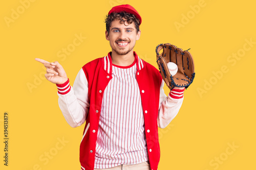 Young handsome man with curly hair wearing baseball uniform holding golve and ball smiling happy pointing with hand and finger to the side photo