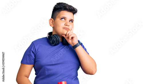 Little boy kid holding skate and wearing headphones serious face thinking about question with hand on chin, thoughtful about confusing idea