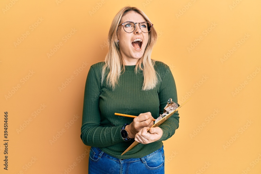 Young caucasian woman writing on checklist clipboard angry and mad screaming frustrated and furious, shouting with anger looking up. Young caucasian woman writing on checklist clipboard angry and mad screaming frustrated and furious, shouting with anger looking up.