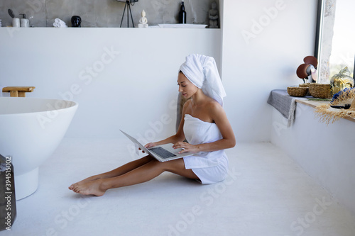 Woman in white towel sits in the bathroom and use a laptop.