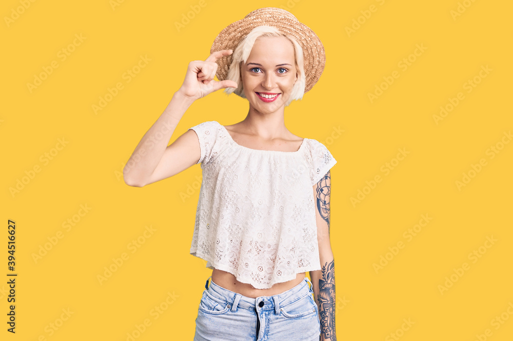 Young blonde woman with tattoo wearing summer hat smiling and confident gesturing with hand doing small size sign with fingers looking and the camera. measure concept. Young blonde woman with tattoo wearing summer hat smiling and confident gesturing with hand doing small size sign with fingers looking and the camera. measure concept.