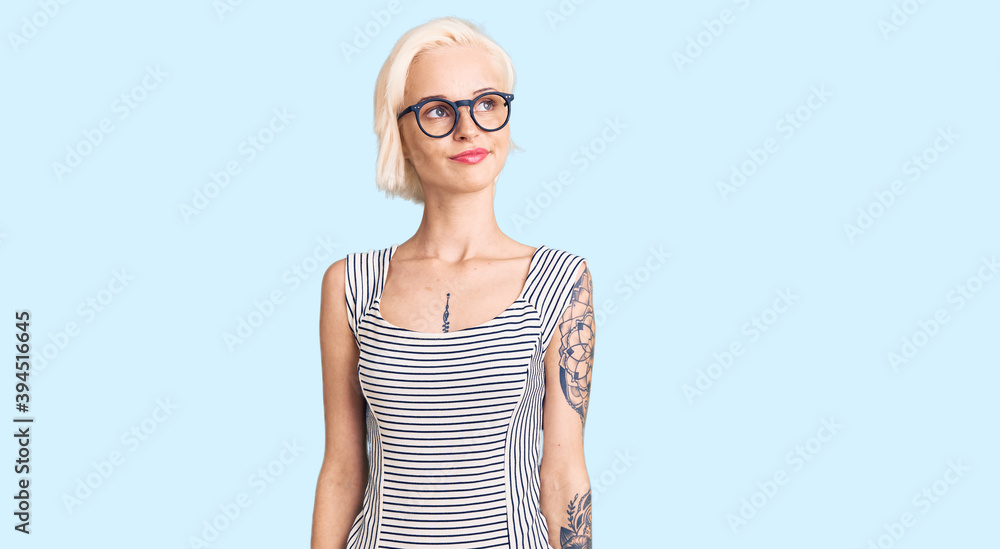Young blonde woman with tattoo wearing casual clothes and glasses smiling looking to the side and staring away thinking. Young blonde woman with tattoo wearing casual clothes and glasses smiling looking to the side and staring away thinking.