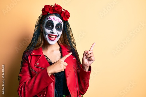 Woman wearing day of the dead costume over yellow smiling and looking at the camera pointing with two hands and fingers to the side.