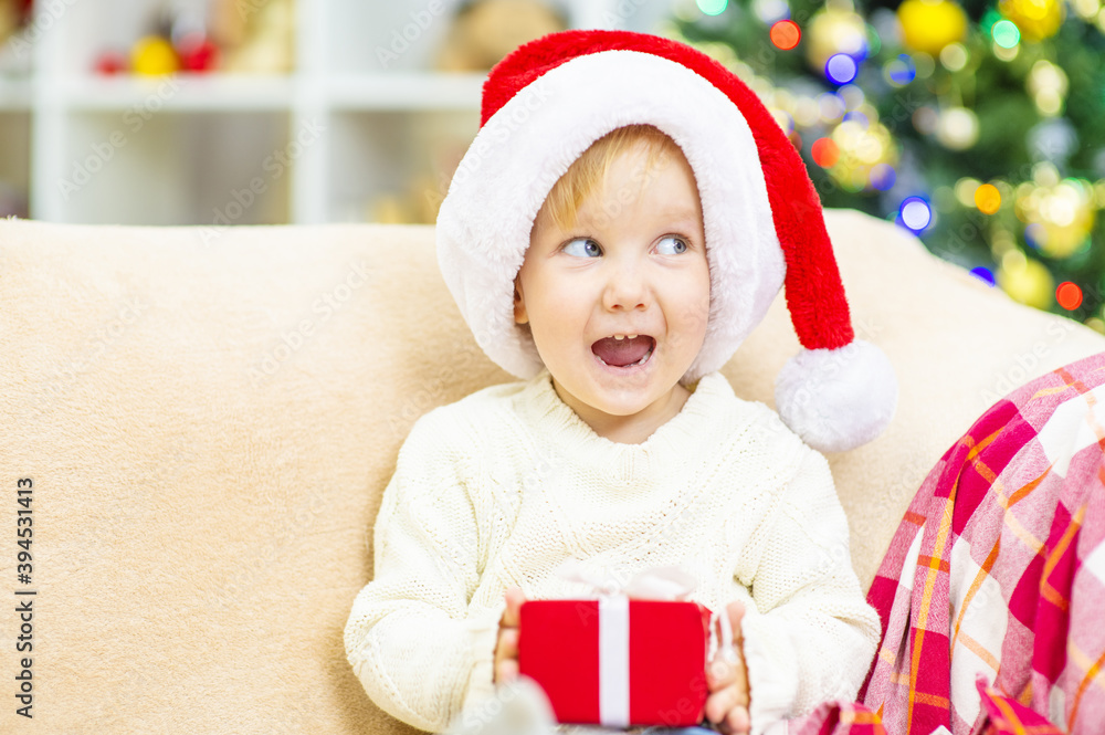 A little girl sits in a Santa hat at home on the sofa on a Christmas tree and holds a gift in her hands. A little girl sits in a Santa hat at home on the sofa on a Christmas tree and holds a gift in her hands.