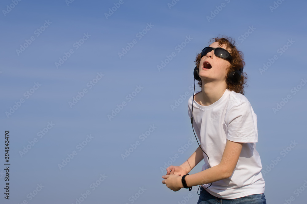 Empty space for text, logo, ad. Teenage boy in headphones and sunglasses is listening to music, dancing and singing outdoors. Empty space for text, logo, ad. Teenage boy in headphones and sunglasses is listening to music, dancing and singing outdoors.