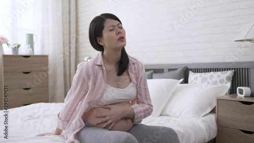 portrait asian woman in her last trimester is groaning and holding her belly while experiencing contraction pain by bedside in the bedroom.